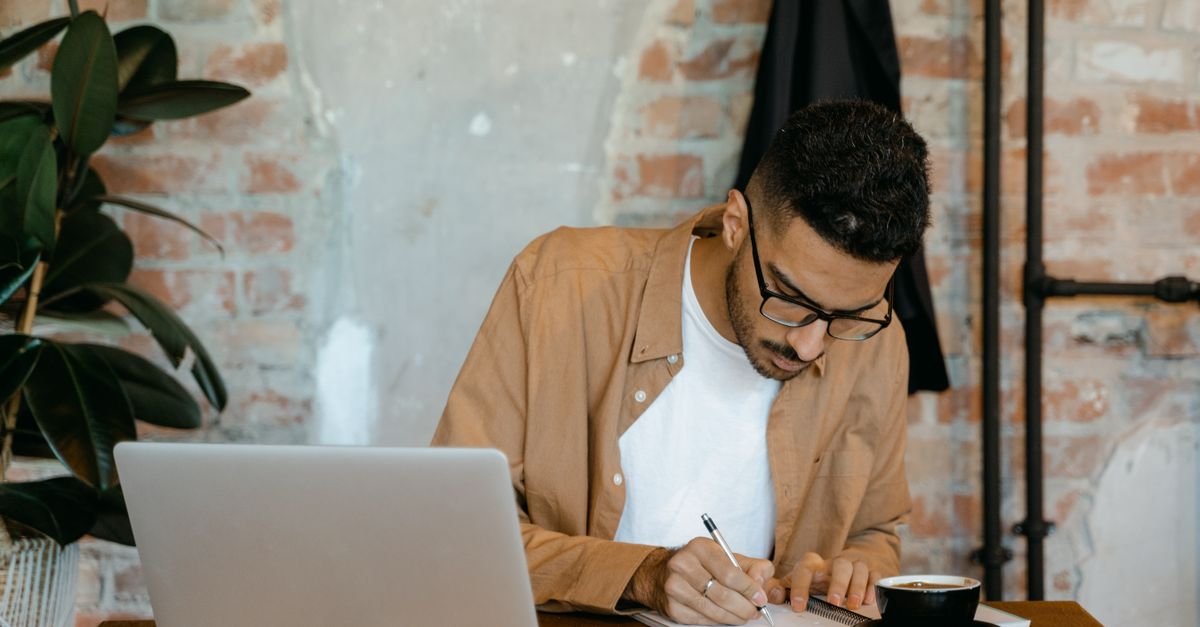 A man writes notes beside his laptop at a home desk, comparing smith ai jobs details.