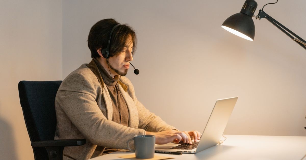 A man wearing a headset types on a laptop while doing part time ai jobs at a desk.